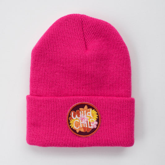 The Wild Child - Fuchsia Kids Beanie by seaslope1 showcases a vibrant pink color and features a cuff design. It is adorned with a colorful embroidered patch on the front, displaying the phrase "wild child" on a simple light gray background.