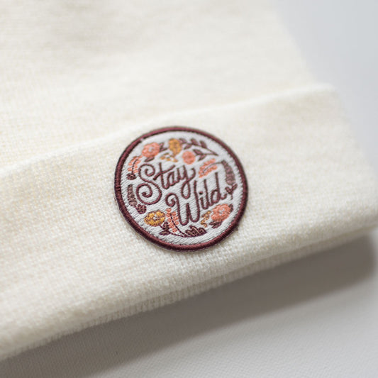 This is a description of a cream-colored knit beanie designed for kids, featuring a round embroidered patch. The patch has the words "Stay Wild" surrounded by vibrantly colored flowers in red, yellow, and pink. Made from 100% acrylic, it offers warmth and style for various outdoor adventures.