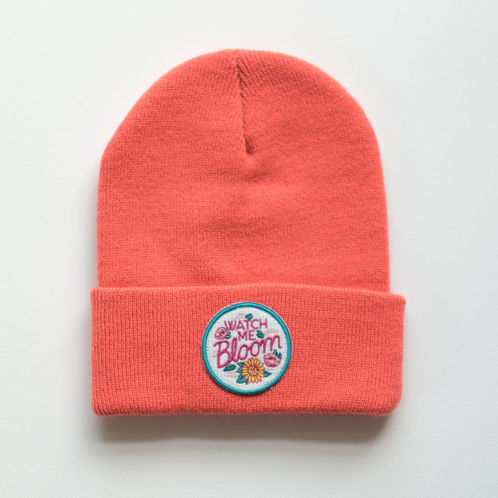 The WATCH ME BLOOM INFANT/TODDLER BEANIE is a coral-colored knit hat made from 100% acrylic. It features a folded brim and an embroidered patch with the phrase "Watch Me Bloom" alongside a cute flower design on a white background.