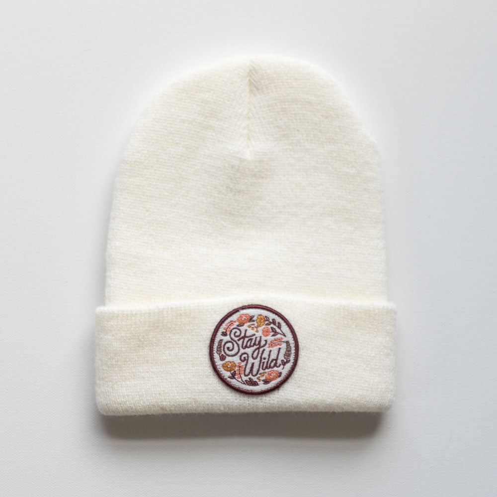 The "Stay Wild - Dove Kids Beanie" by seaslope is a stylish white knit hat made from 100% acrylic. It features an embroidered patch on the front with the words "Stay Wild" surrounded by delicate floral designs.