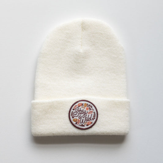 The "Stay Wild - Dove Kids Beanie" by seaslope is a stylish white knit hat made from 100% acrylic. It features an embroidered patch on the front with the words "Stay Wild" surrounded by delicate floral designs.