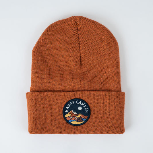 This burnt orange knit beanie, dubbed "Happy Camper | Canyon - Adult Beanie" by seaslope1, is adorned with a circular patch featuring mountain and moon motifs. Displayed on a neutral backdrop, it showcases the allure of handmade artistry.