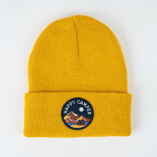 The Happy Camper - Sun Kids Beanie is a stylish mustard yellow knit hat that features a prominent knit texture and a folded brim. It includes an embroidered patch in the center labeled "HAPPY CAMPER," showcasing mountains and a moon, making it perfect for children.