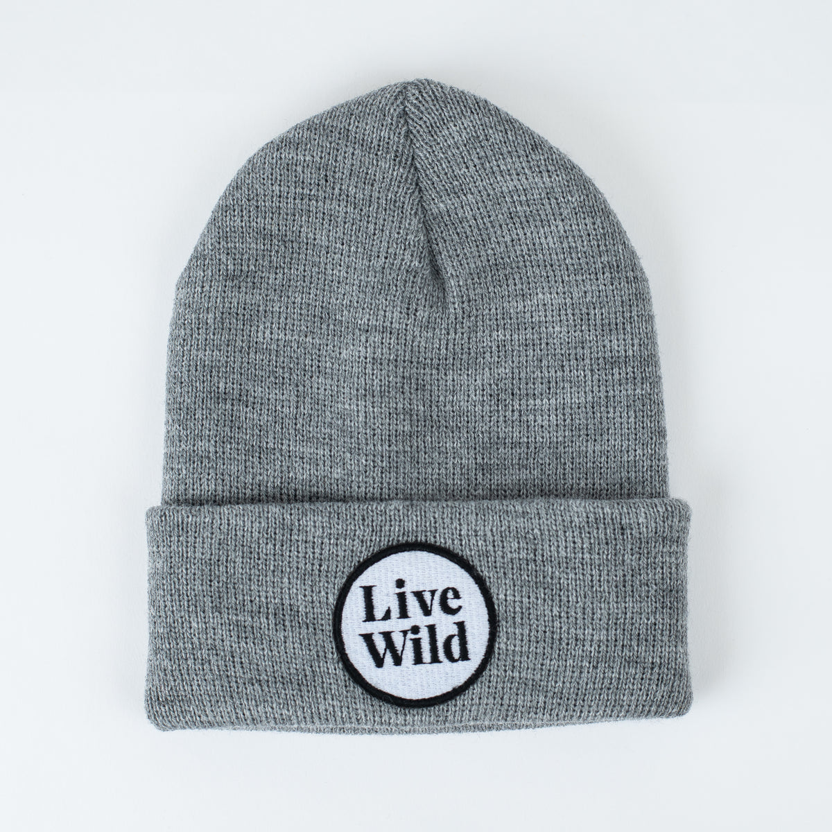 The "Live Wild - Stone Kids Beanie" is a gray knit beanie with a folded brim. It features an embroidered circular patch that displays the phrase "Live Wild" in black text on a white background.