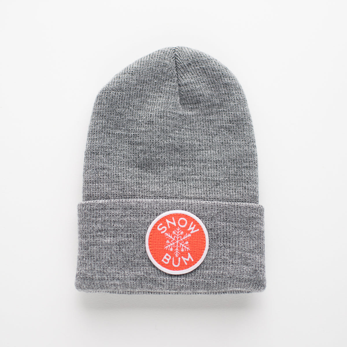 The Snow Bum - Stone Kids Beanie is a gray beanie with a folded cuff, made from 100% acrylic. It features an orange circular embroidered patch displaying a white snowflake and the words "SNOW BUM" in white letters. The product is shown against a plain white background.