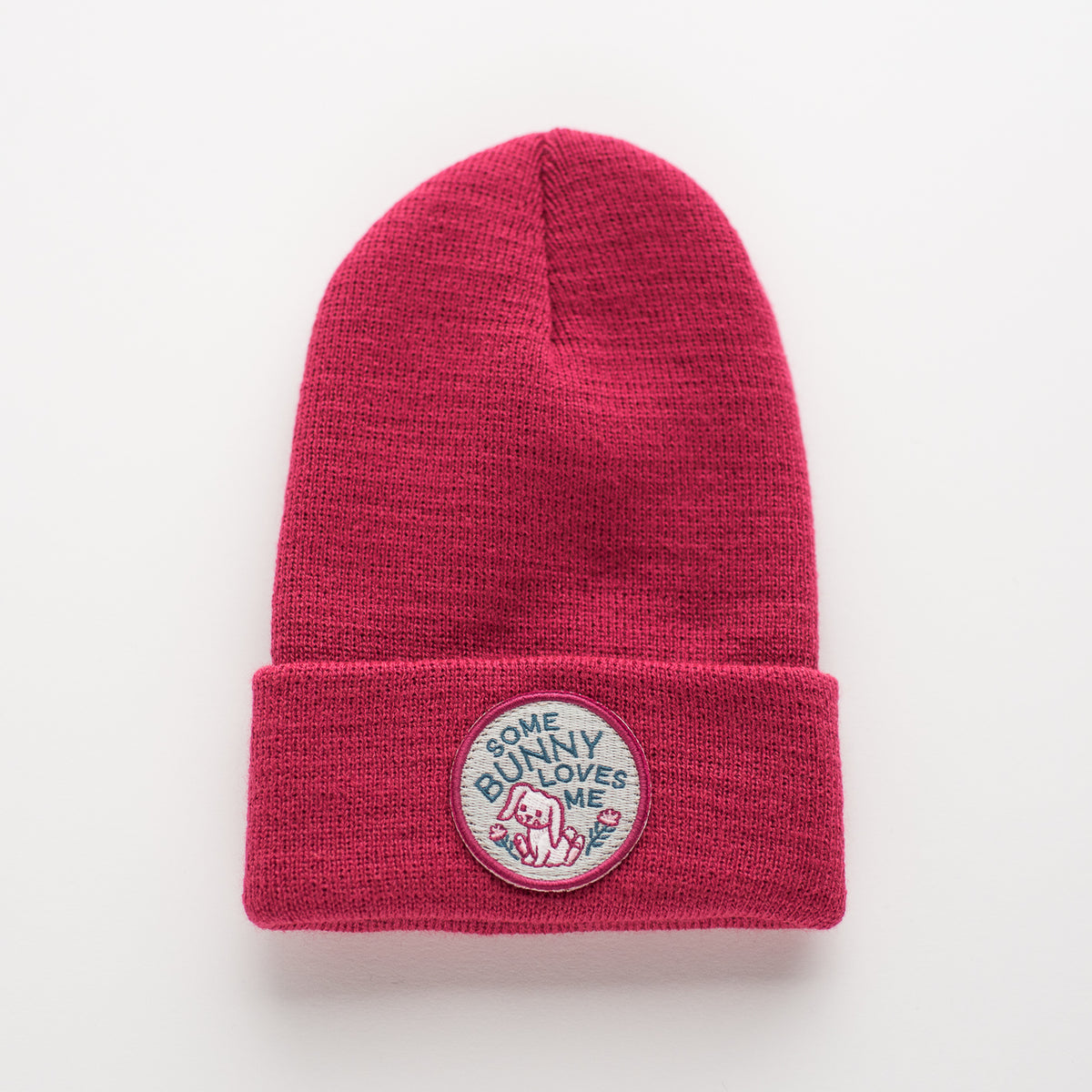 Introducing the "Some Bunny - Tulip Kids Beanie," a charming red knit beanie crafted from 100% acrylic. This cozy hat features a folded brim, accentuated by an embroidered circular patch that displays an adorable bunny design with the phrase "Some bunny loves me.