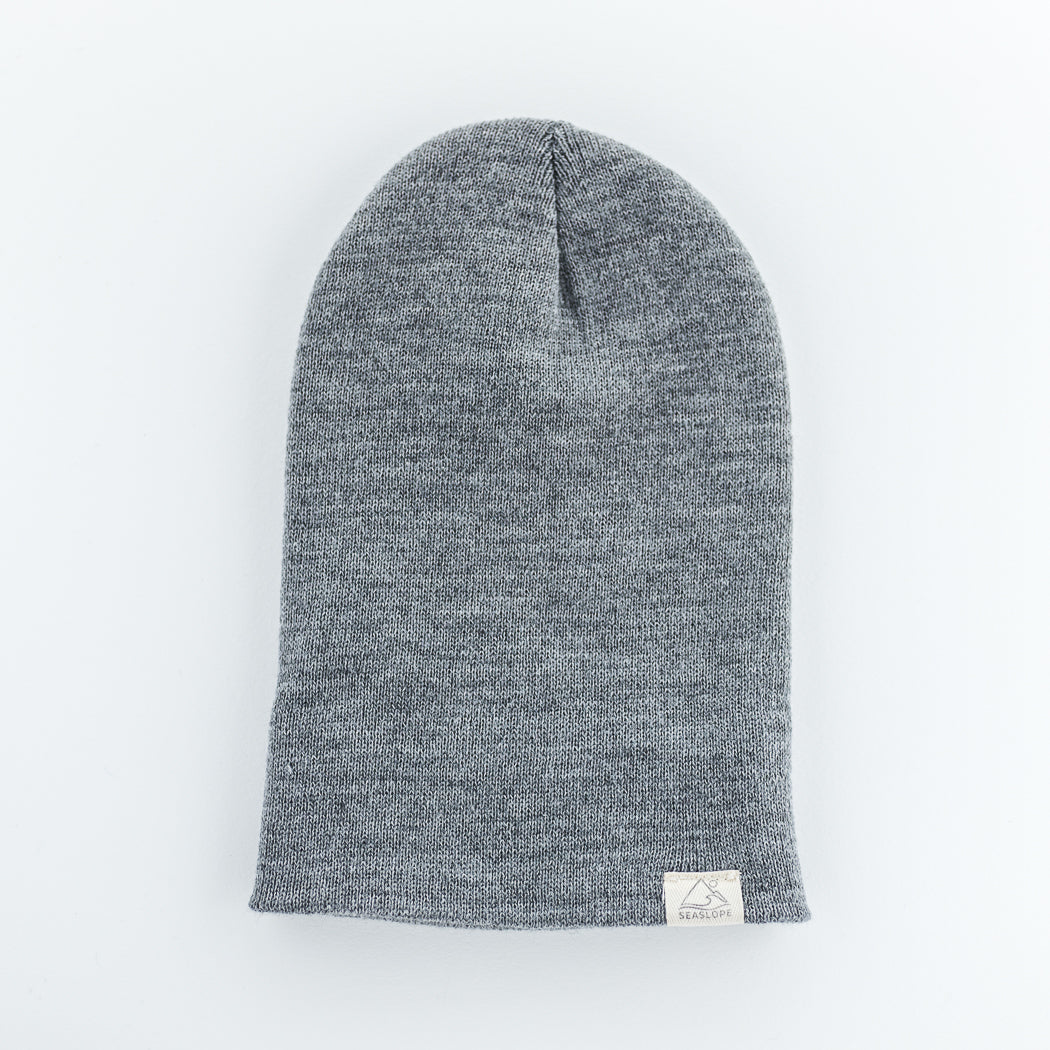 The STONE Youth/Adult Beanie by Seaslope1 is showcased on a light background, emphasizing its soft, 100% acrylic textured fabric. This gray knit beanie, made in the USA, includes a small white label with a minimalist design at the bottom.