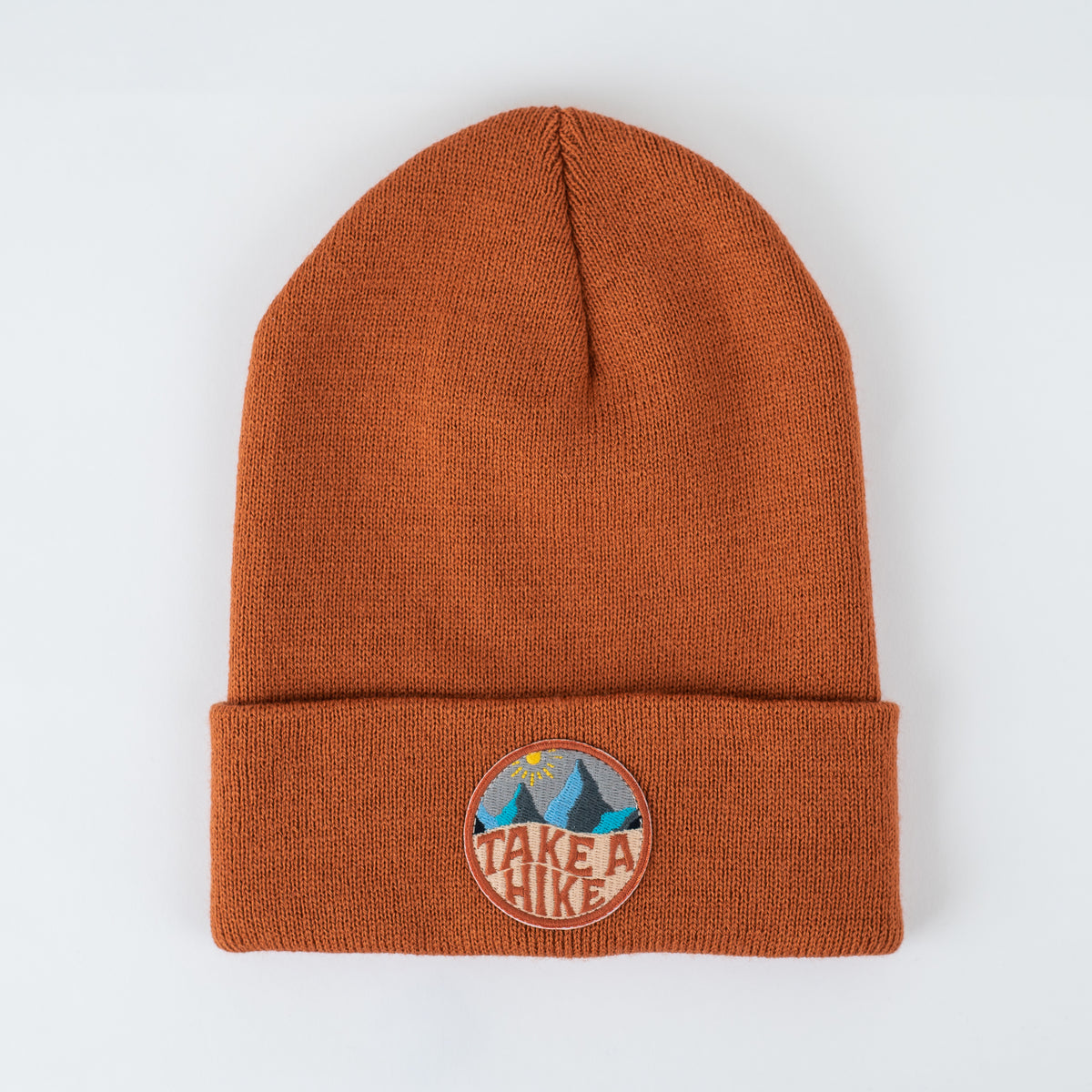 Rust knit beanie with a folded brim and a round patch depicting blue mountains and a yellow sun; cozy, handmade style.