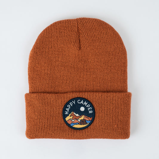 The seaslope1 Happy Camper - Canyon Kids Beanie is a rust-colored, acrylic beanie featuring a folded brim and a round patch. The patch depicts a mountain scene under a crescent moon with "HAPPY CAMPER" embroidered above it. This beanie is made in the USA.