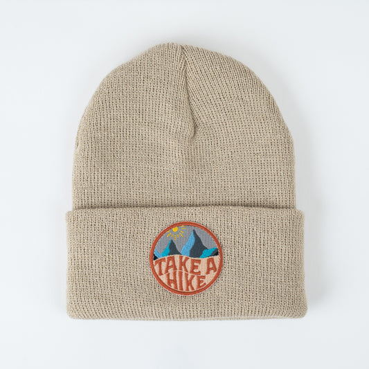 Introducing the seaslope1 TAKE A HIKE SAND INFANT/TODDLER BEANIE, a soft stretch fit beige knit design featuring a circular embroidered patch. The patch showcases a mountain motif with the sun and the bold phrase "Take a Hike." Proudly made in the USA, it’s an ideal choice for little adventurers seeking both style and comfort.