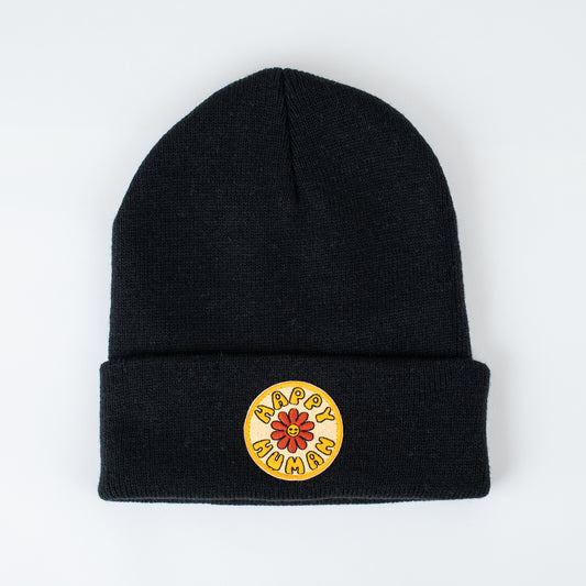 The adult beanie features a black knit design with a folded edge, highlighted by a circular patch. The yellow patch includes red and black text that reads "Happy Human," adorned with a central red flower. This handcrafted item is presented on a plain white background.