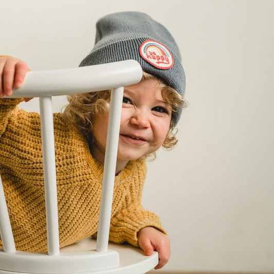 A young child with curly hair smiles while leaning on a white chair, wearing a mustard sweater and a gray beanie. The soft and stretchy fit guarantees comfort, with the 100% acrylic material standing out against the light-colored background.