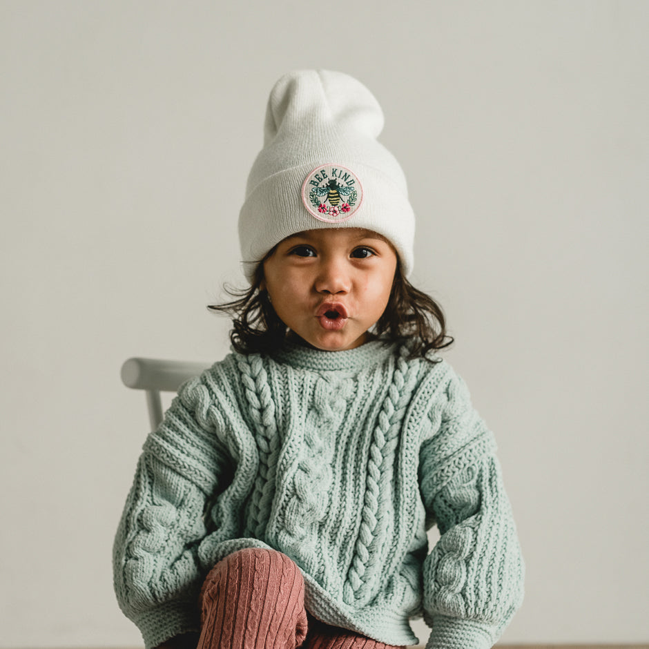 A child with long hair sits on a chair, wearing a mint green cable-knit sweater and a "Bee Kind - White Kids Beanie," proudly displaying its "Bee Kind" patch. Their animated expression with puckered lips adds charm against the plain background.