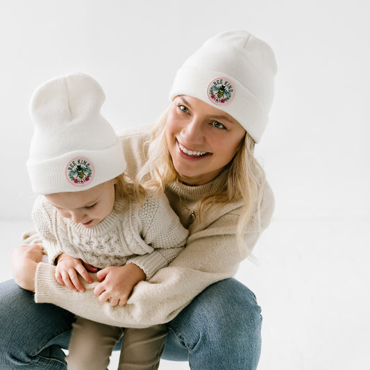 A woman and a toddler, both wearing matching white beanies, share a tender moment. The woman smiles warmly while holding the toddler, who looks down. They are dressed in light sweaters against a white backdrop, and their charming hats complete the scene. Proudly made in the USA with love!