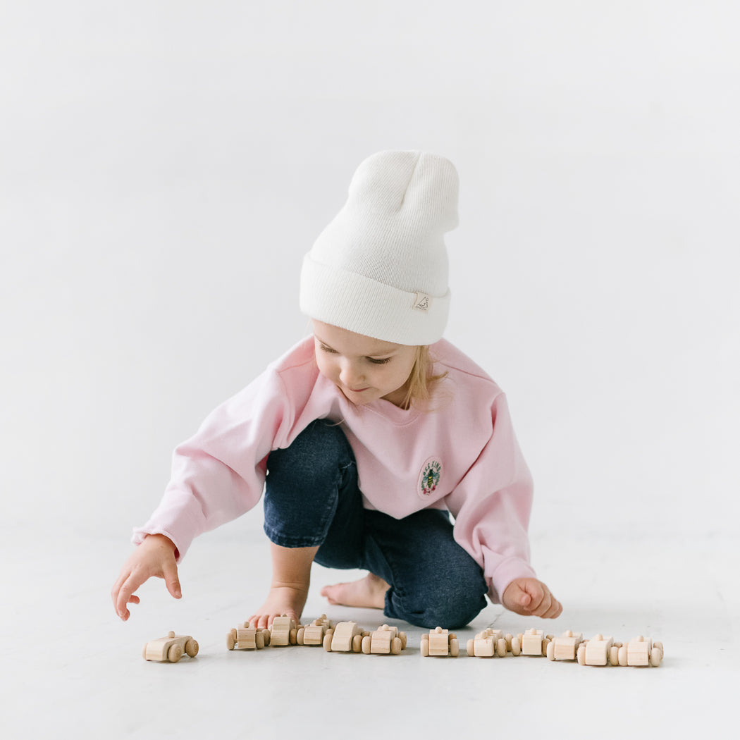 A young child in a pink sweater crouches on the floor, playing with wooden toy cars arranged in a line. They are wearing a cozy beanie, which adds to the warmth of the scene set against a plain, white background.