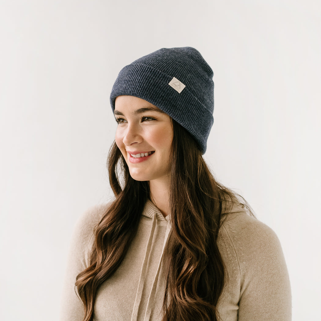 A person with long brown hair smiles while looking off to the side, wearing a Dusk Youth/Adult Beanie paired with a beige hooded sweater. The plain, light-colored background complements their relaxed vibe.