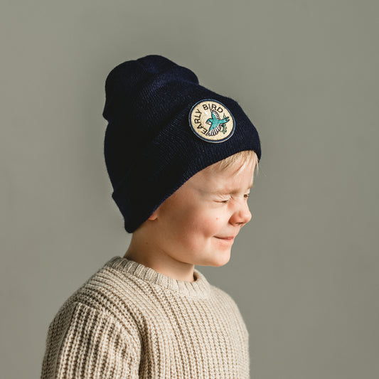 A young child in a beige knit sweater smiles with eyes closed, proudly wearing a navy kids beanie. The dark blue beanie, adorned with an "Early Bird" patch featuring a bird illustration on the side, complements the neutral gray background perfectly.