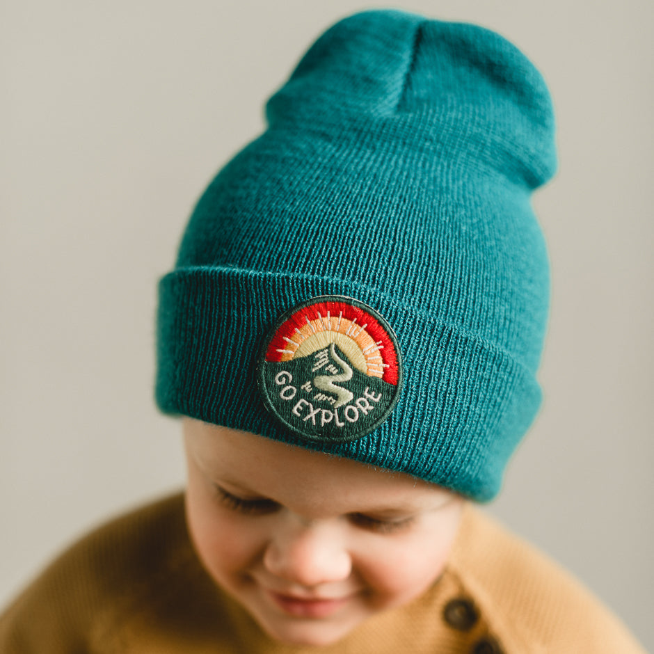 A baby wearing a teal knit beanie beams with joy while looking downward. The beanie, featuring an embroidered patch, is made entirely of acrylic and complements the baby's brown sweater beautifully against the light, neutral background.