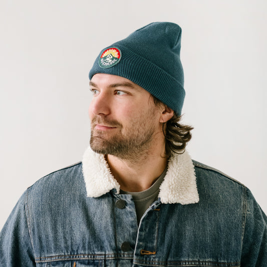 A person with a beard wears a stylish blue beanie, adorned with a colorful patch featuring a mountain and sun design, adding flair to his look. He complements it with a denim jacket that has a shearling collar as he looks to the side against a plain, light-colored background.