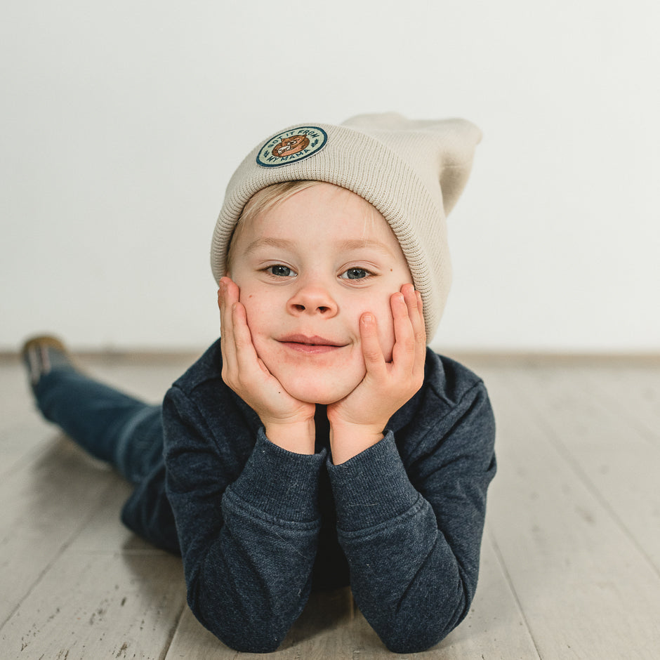 A young child lies on their stomach on a wooden floor, wearing a sand-colored beanie with an embroidered patch. Dressed in a dark blue long-sleeve shirt, they rest their chin on their hands and smile warmly at the camera.