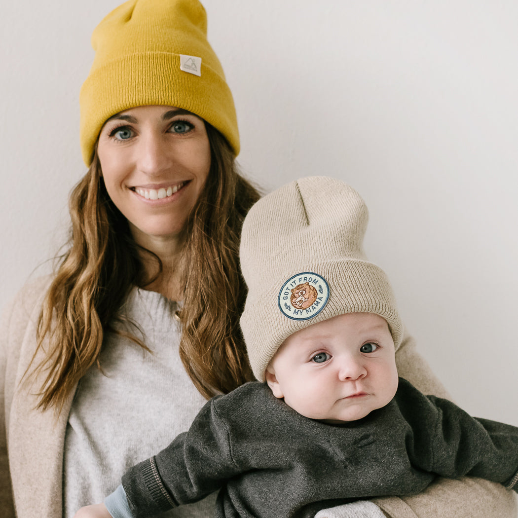 A woman with a warm smile is wearing a yellow knit beanie and holding a baby. The baby is also wearing a beanie, specifically the "Got it from my Mama - Sand Kids Beanie." They are both standing against a plain white background, looking directly at the camera.