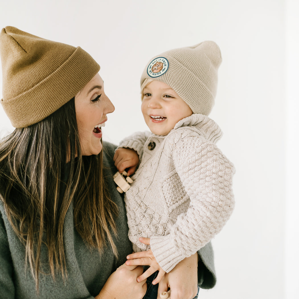 A woman and a child share a joyful moment, smiling at each other while dressed in cozy sweaters and knit beanies. The child's beanie includes an embroidered patch and is part of the "Got it from my Mama - Sand Kids Beanie" collection. The scene is set against a simple, light background as the child is held lovingly in the woman's arms.