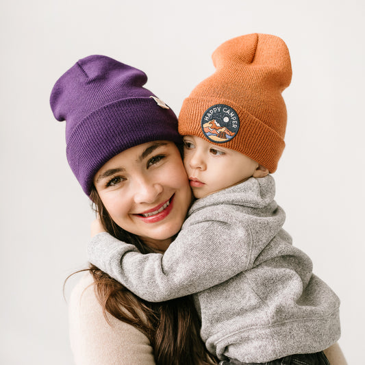 A woman and child are posing together, wearing vibrant knit beanies. The woman is wearing a purple beanie and smiling as she holds the child. The child, leaning affectionately on her shoulder, is wearing an orange beanie with a cheerful patch.