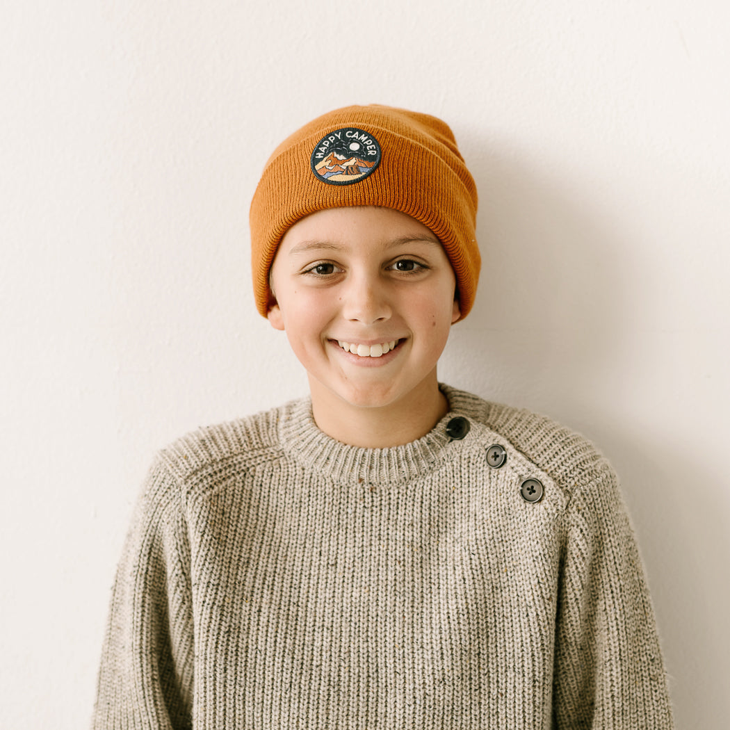 A smiling child wearing a burnt orange knit beanie with a graphic patch and a gray sweater featuring button details on the shoulder stands against a plain white background.