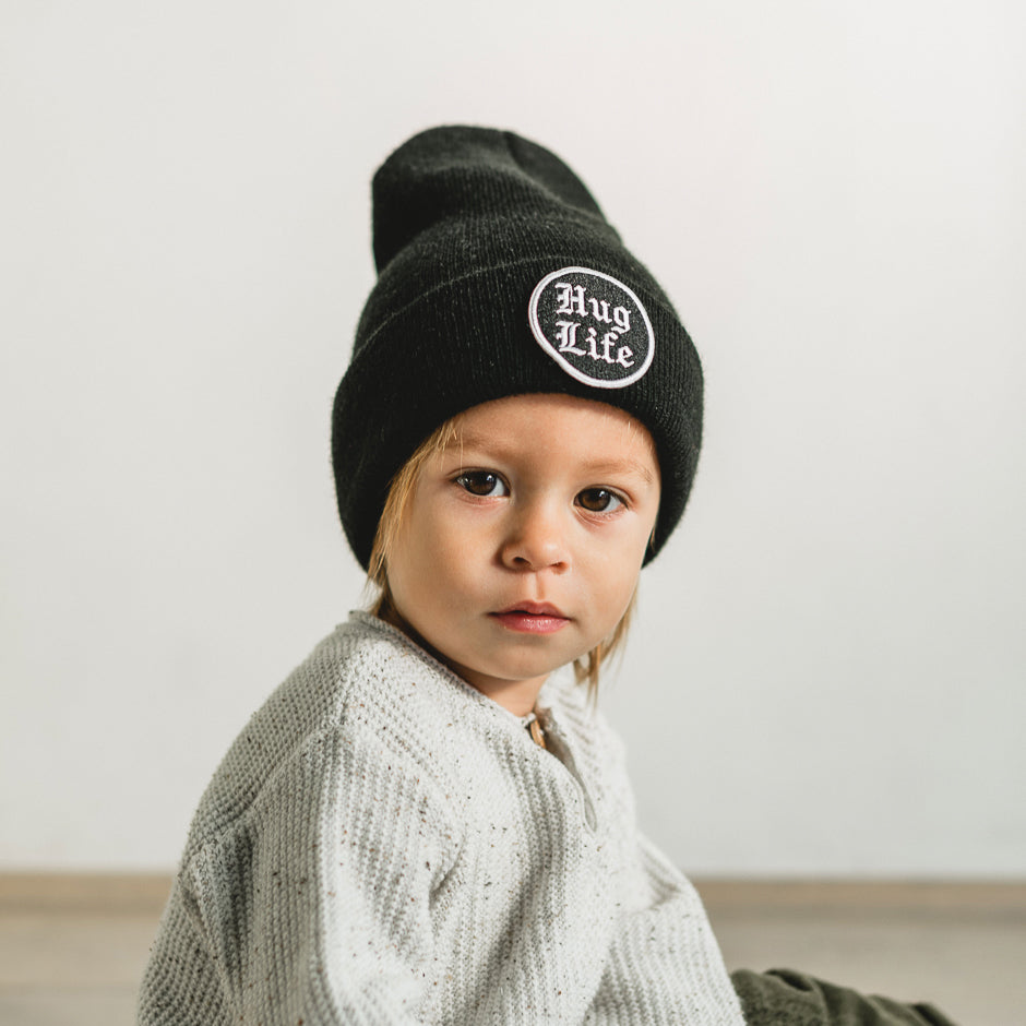 A young child sits on the floor, looking intently at the camera, wearing a jet black beanie with an embroidered patch. The background is a plain, light-colored wall.