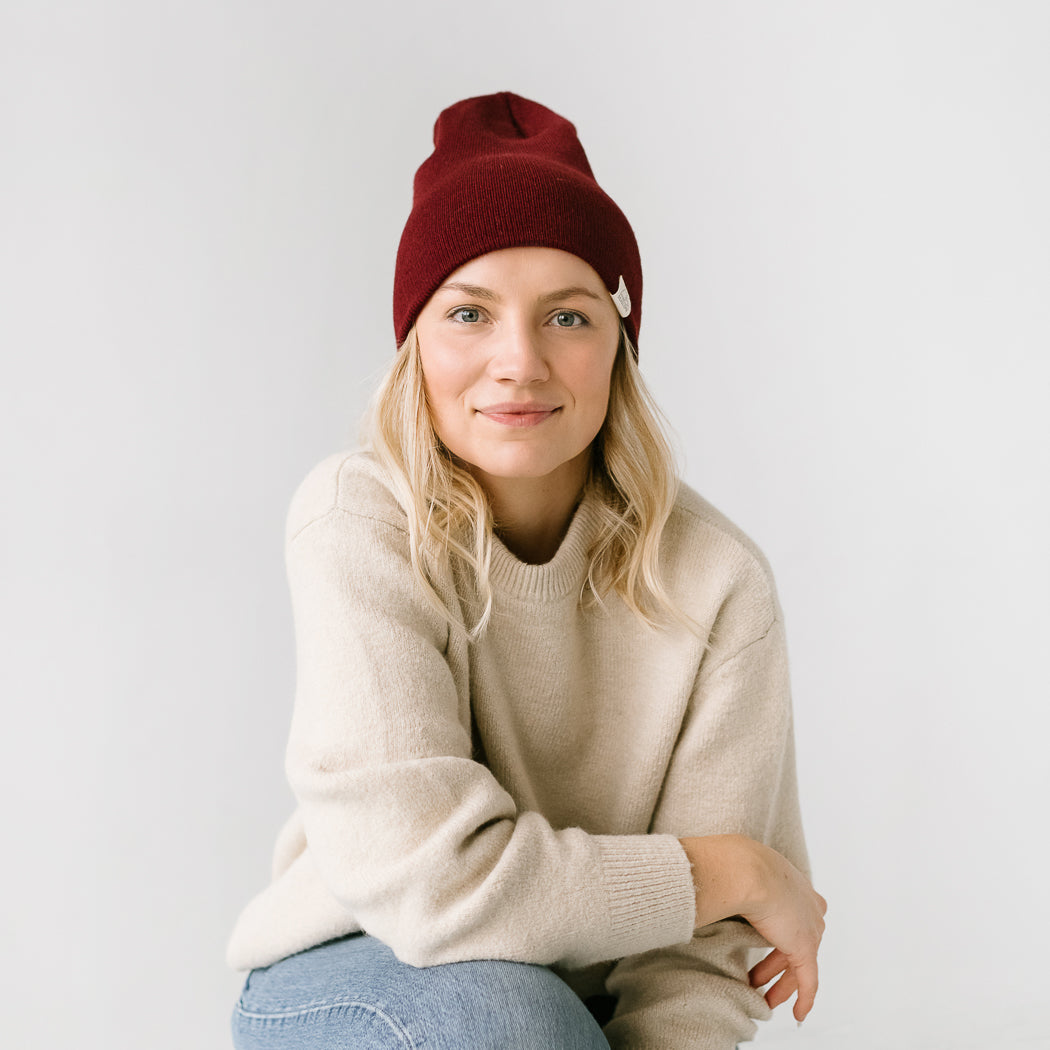 A person wearing a seaslope1 Maple youth/adult beanie, along with a beige sweater and blue jeans, sits against a plain white background while smiling gently.