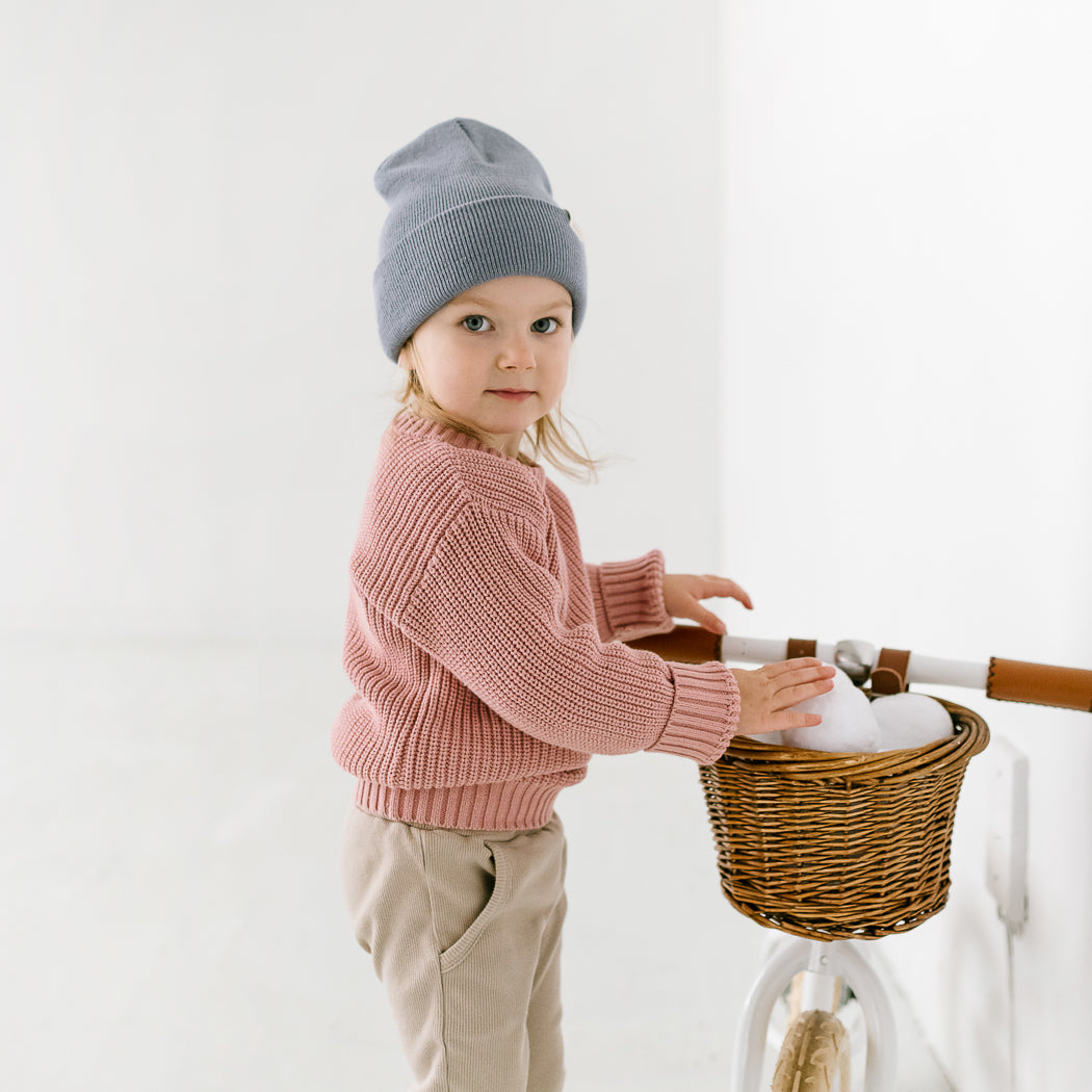 A young child in a pink sweater is wearing a beanie, standing next to a bicycle with a wicker basket filled with snowballs. The scene is set against a minimalist white background.