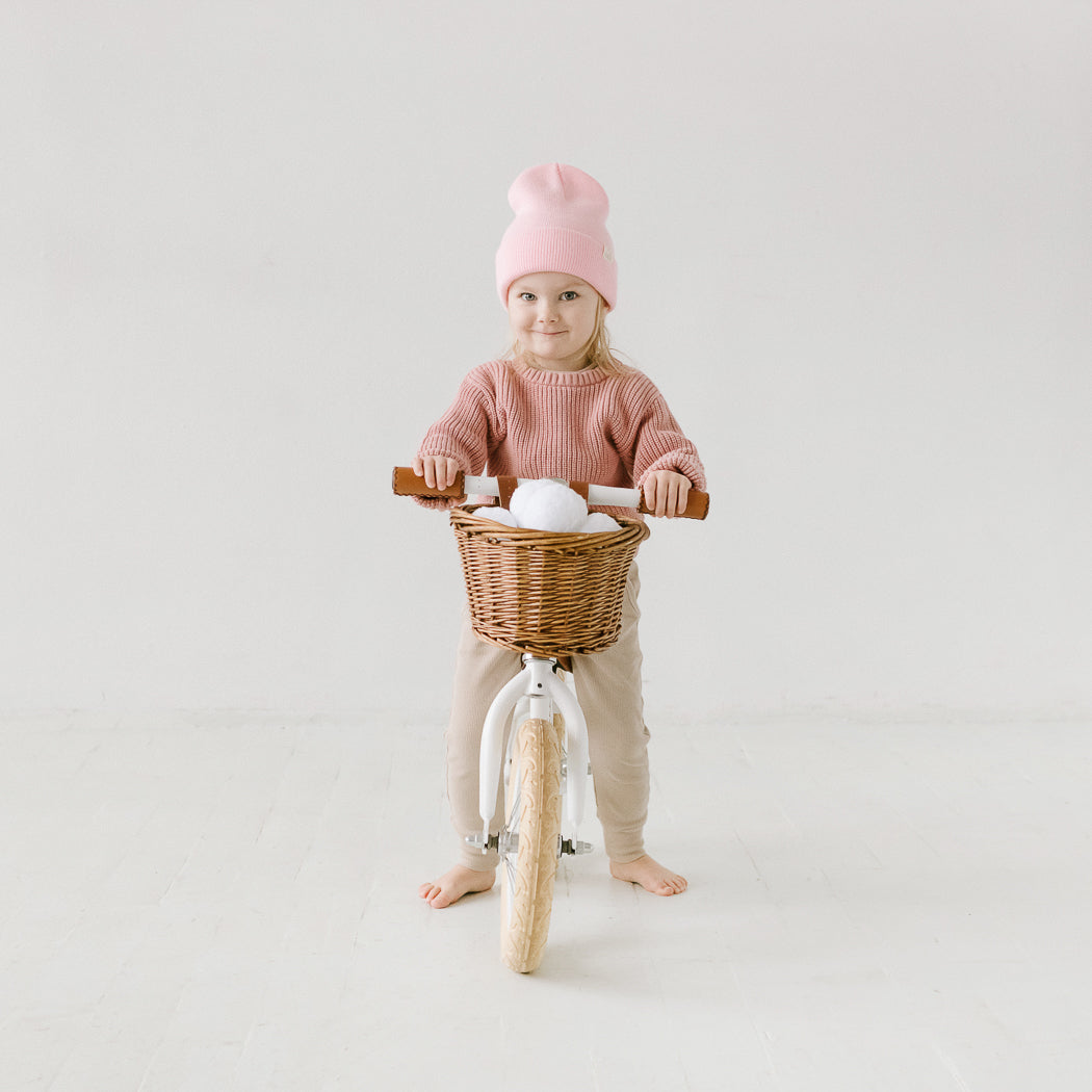 A young child is wearing a pink beanie and sweater, standing barefoot next to a white balance bike. The bike has a wicker basket that contains a stuffed toy. The scene is set against a plain, light-colored wall with accents made in the USA.