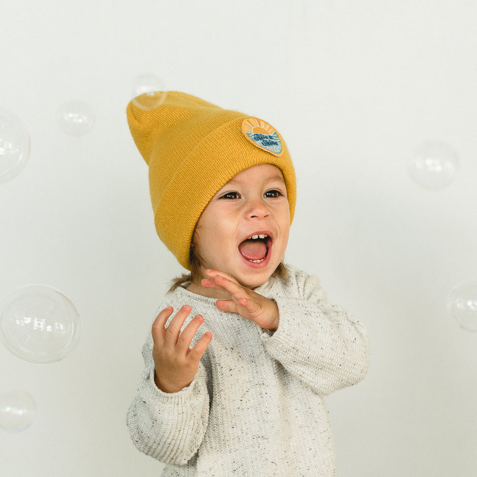 A joyful child wearing a colorful beanie and a gray sweater is surrounded by floating bubbles. The child looks up with a big smile and open mouth, clearly delighted by the bubbles against a plain white background.