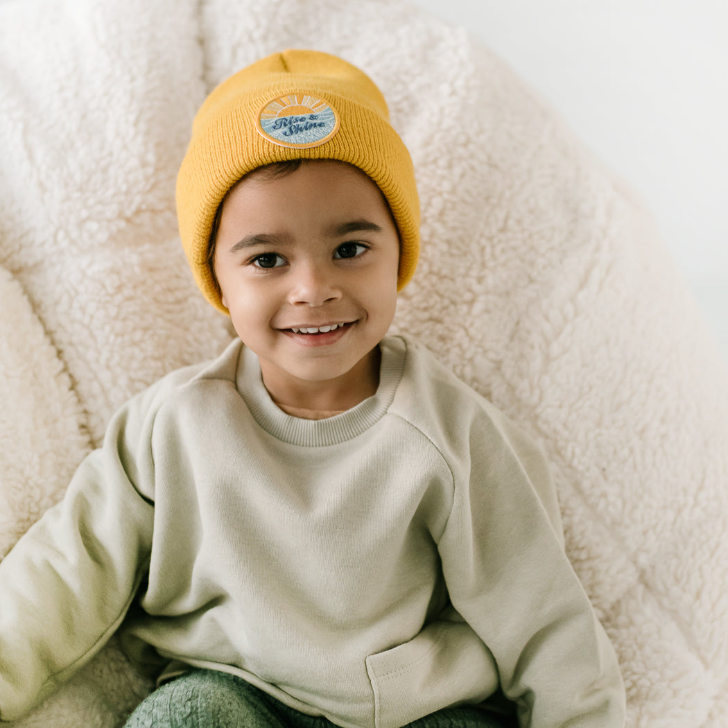 A young child wearing a beanie sits on a fluffy cream-colored chair, while donning a light beige sweater. Smiling and looking at the camera, the child creates a warm and cheerful atmosphere.