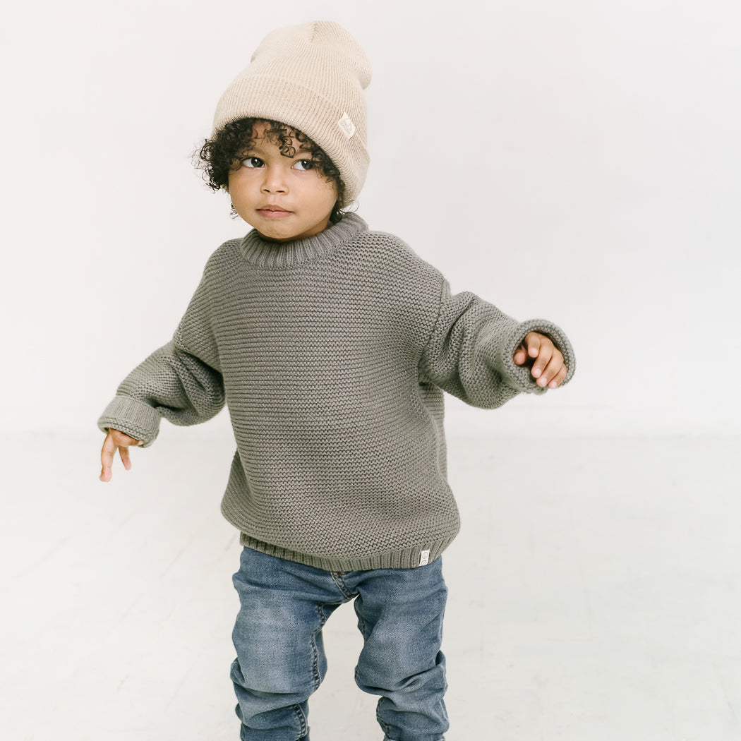 A young child with curly hair is dressed in a cozy knit beanie, a gray oversized sweater, and blue jeans. They stand against a light background, and the soft stretch fit of the beanie enhances their expression of curiosity.