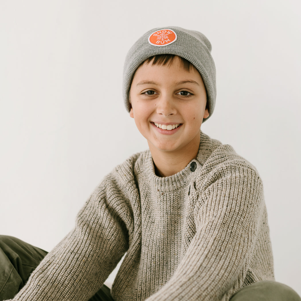 A smiling person in a gray handmade knit sweater and an adult beanie sits against a plain white background, gazing warmly at the camera.