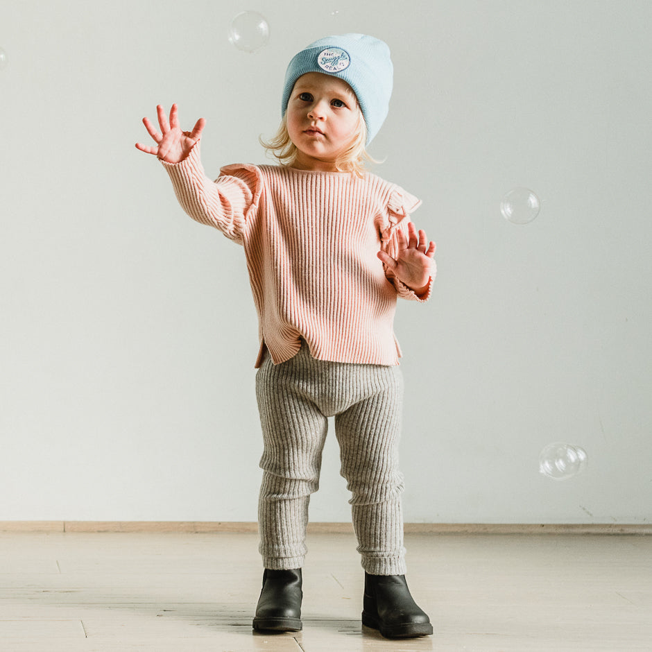 A toddler wearing a cozy beanie, along with a peach sweater, gray leggings, and black boots, stands indoors on a wooden floor. The child has a focused expression as they reach out towards floating bubbles.