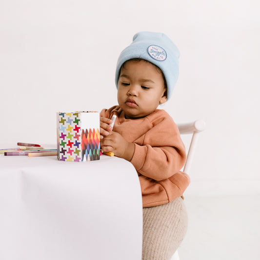 A toddler in a peach sweatshirt is seated at a table, holding a yellow crayon. Nearby, a cup with colorful plus signs contains additional crayons. The child appears focused on drawing, adding charm to the cozy scene with their embroidered patch-adorned hat.