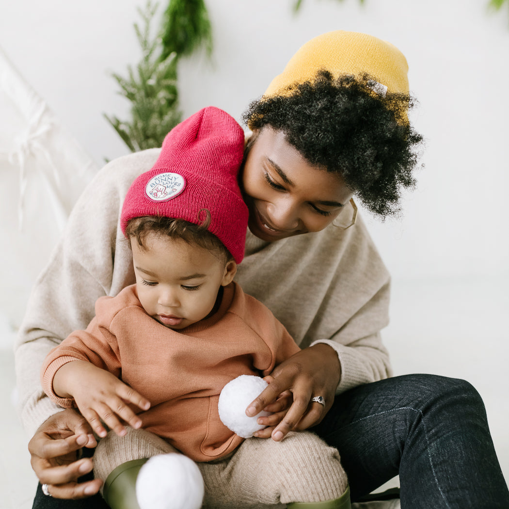 A person and a child, both sporting vibrant beanies, sit on a snowy surface. The person smiles warmly while holding the child, who is focused on a small snowball in their hands. A festive tree stands in the background, enhancing the cozy scene.