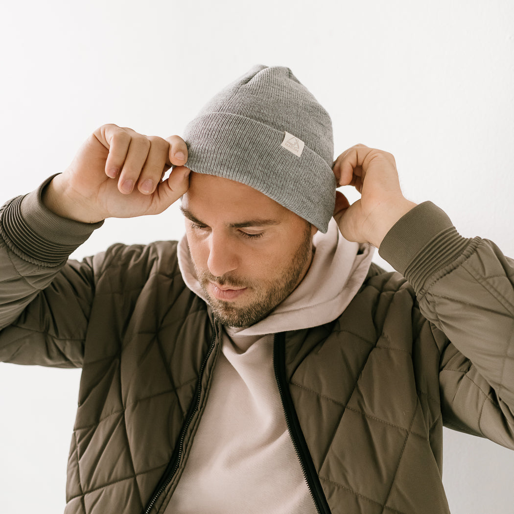 A man is adjusting his beanie, which is made in the USA. He is dressed in a quilted olive green jacket layered over a beige hoodie, standing against a neutral backdrop.