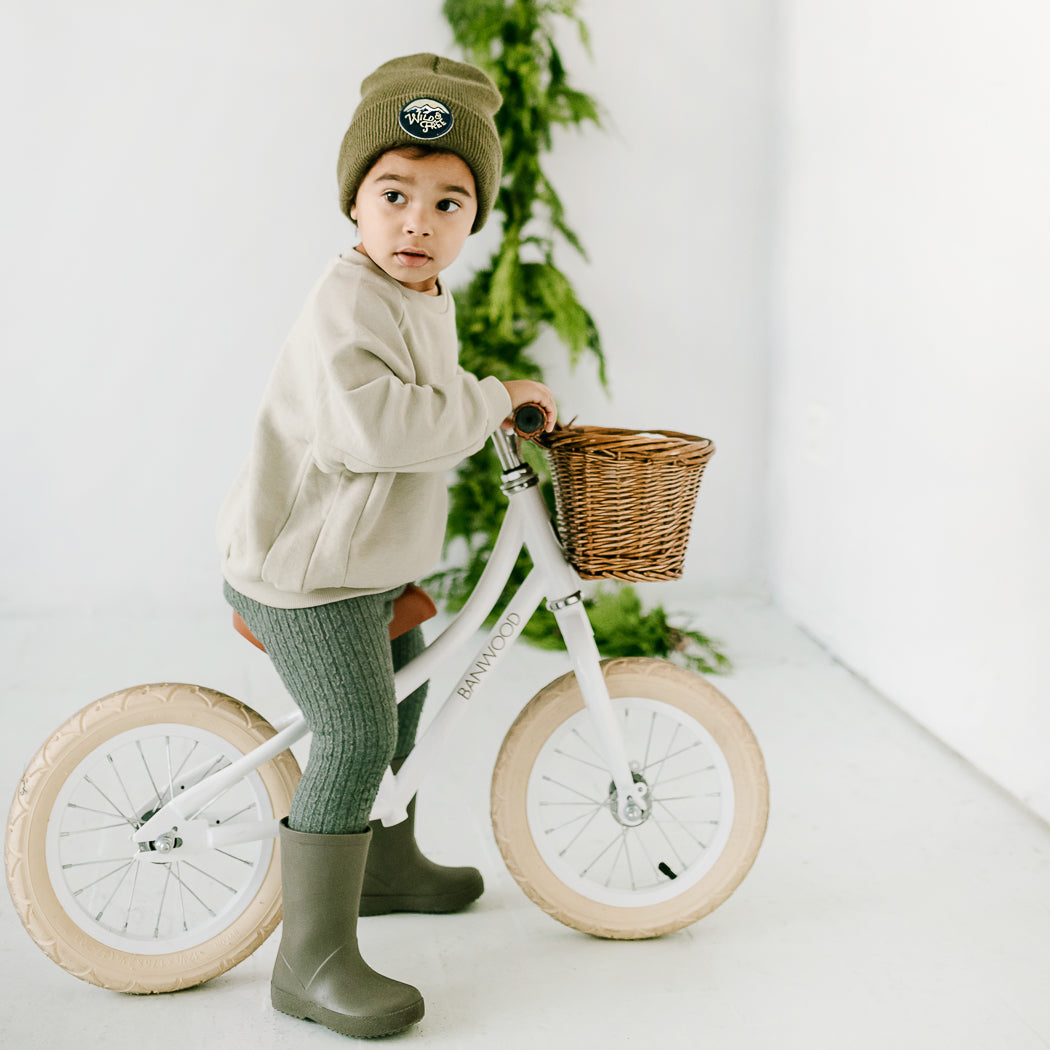 A young child is dressed in a beige sweater, gray leggings, and green boots. They are wearing a beanie and are seated on a white balance bike that has a wicker basket attached. The setting is bright and features green foliage in the background.