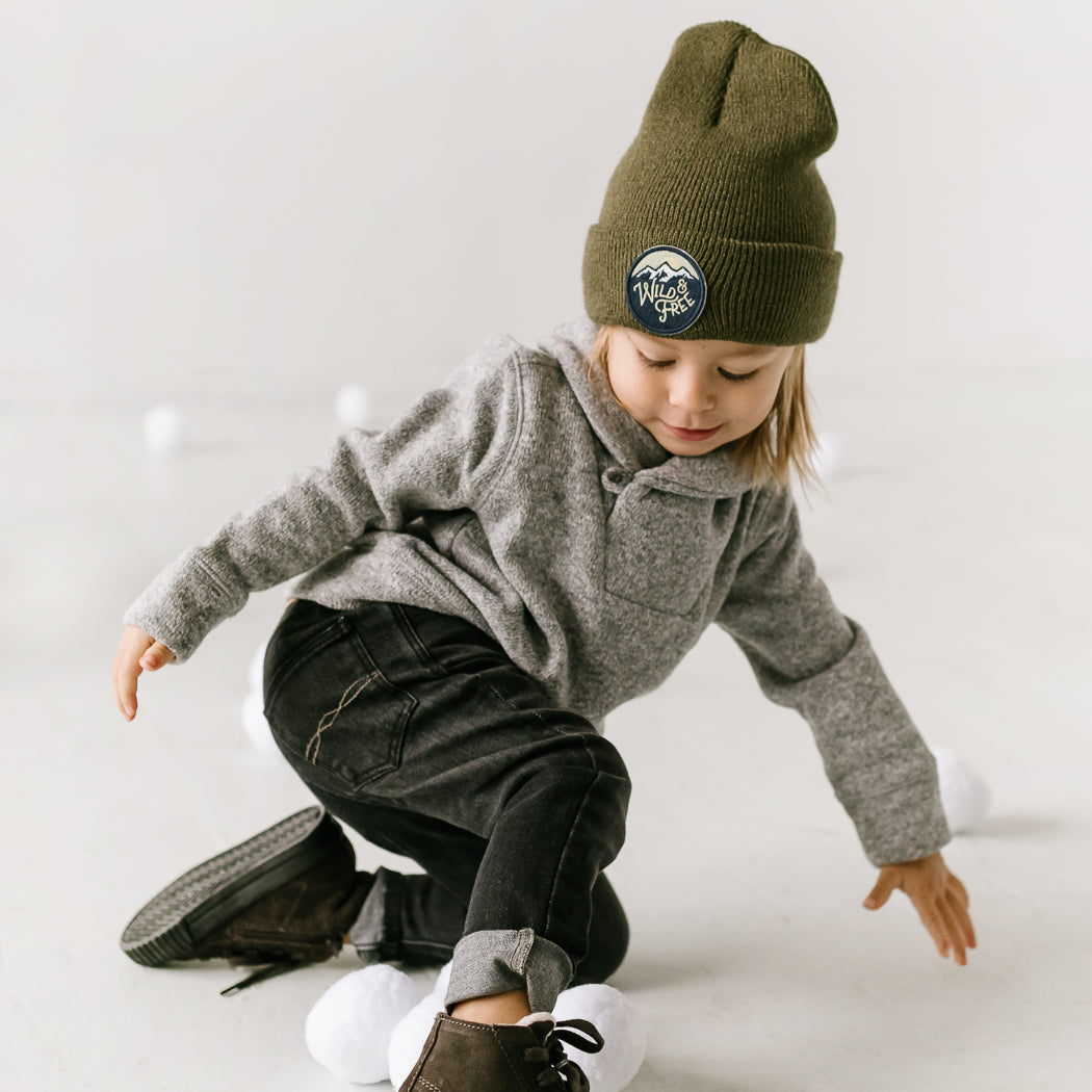 A young child, dressed in a gray sweater, a cozy beanie, and dark jeans, is playfully kneeling on the floor. The scene is filled with soft, white objects that resemble snowballs.